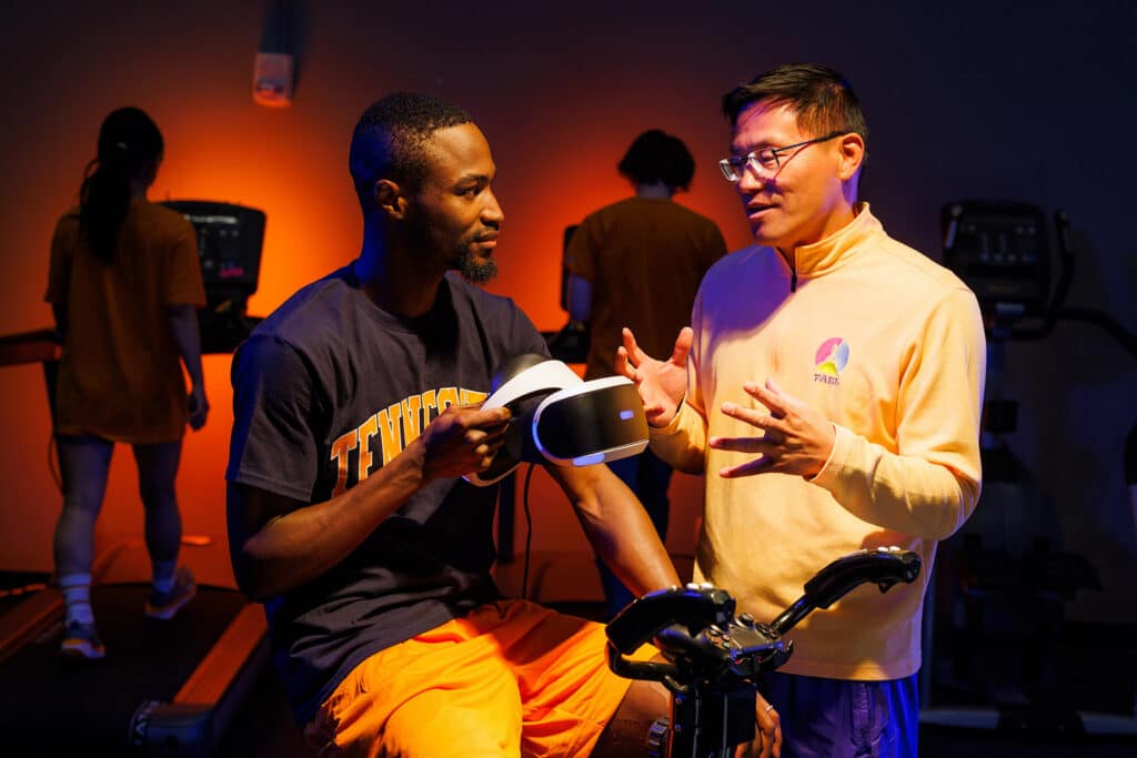 Professor and Department Head Zan Gao talks with kinesiology PhD student John Oginni simulating an experiment using a virtual reality bicycle exercise program in the Physical Activity Epidemiology Lab.